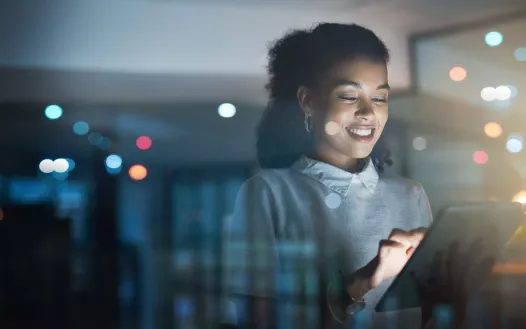 A person stands indoors, smiling while using a tablet. The background is softly blurred with lights.