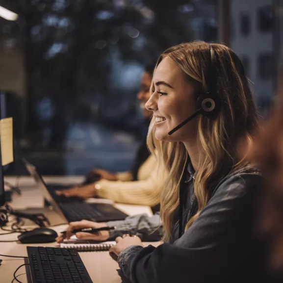 Smiling blond female customer service representative wearing headset using computer at desk in call center.