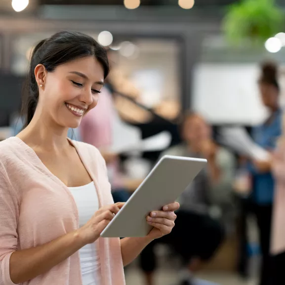 Portrait of a happy woman working online at a creative office using a tablet computer with a group at the background and smiling.