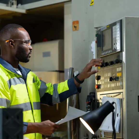 A man in a high-visibility jacket operates a control panel in an industrial setting, holding a sheet of paper.