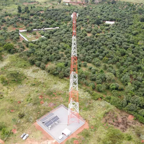 Aerial view of a tall communication tower surrounded by green vegetation, with solar panels and a small building at its base.