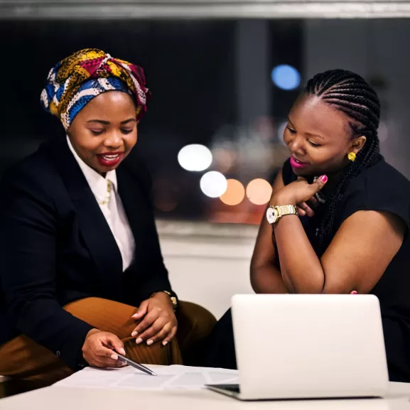 Two women sit at a table, discussing paperwork. One points at the document with a pen. A laptop is open in front of them. The background is blurred and shows city lights through a window.