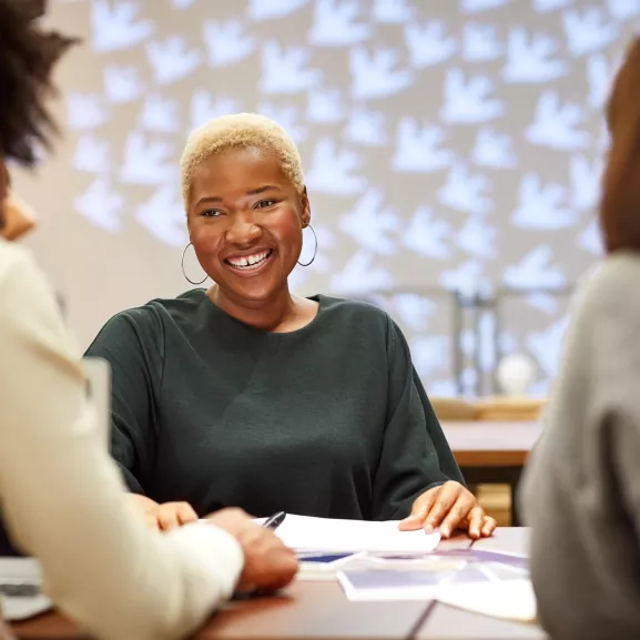 A person with short hair smiling while sitting at a table with others, engaged in discussion.