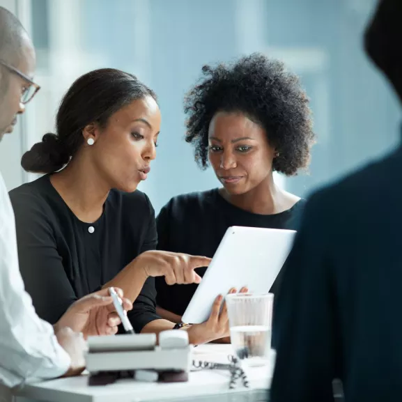 Four people in a meeting, two women focused on a tablet while two others listen.