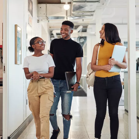 Three people walk down a hallway together, smiling and carrying books and folders.