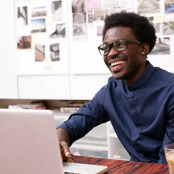 Person in a blue shirt sitting at a desk with a laptop, smiling. A glass of coffee is on the table. A wall with photos and notes is in the background.