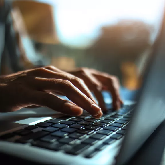 Close-up of hands typing on a laptop keyboard with natural light streaming in from the background.