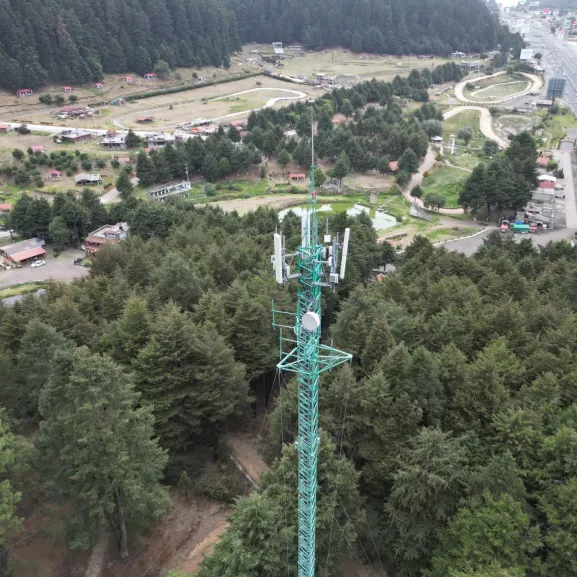 Aerial view of a green telecommunications tower surrounded by dense forest and rural area with roads and buildings in the background.