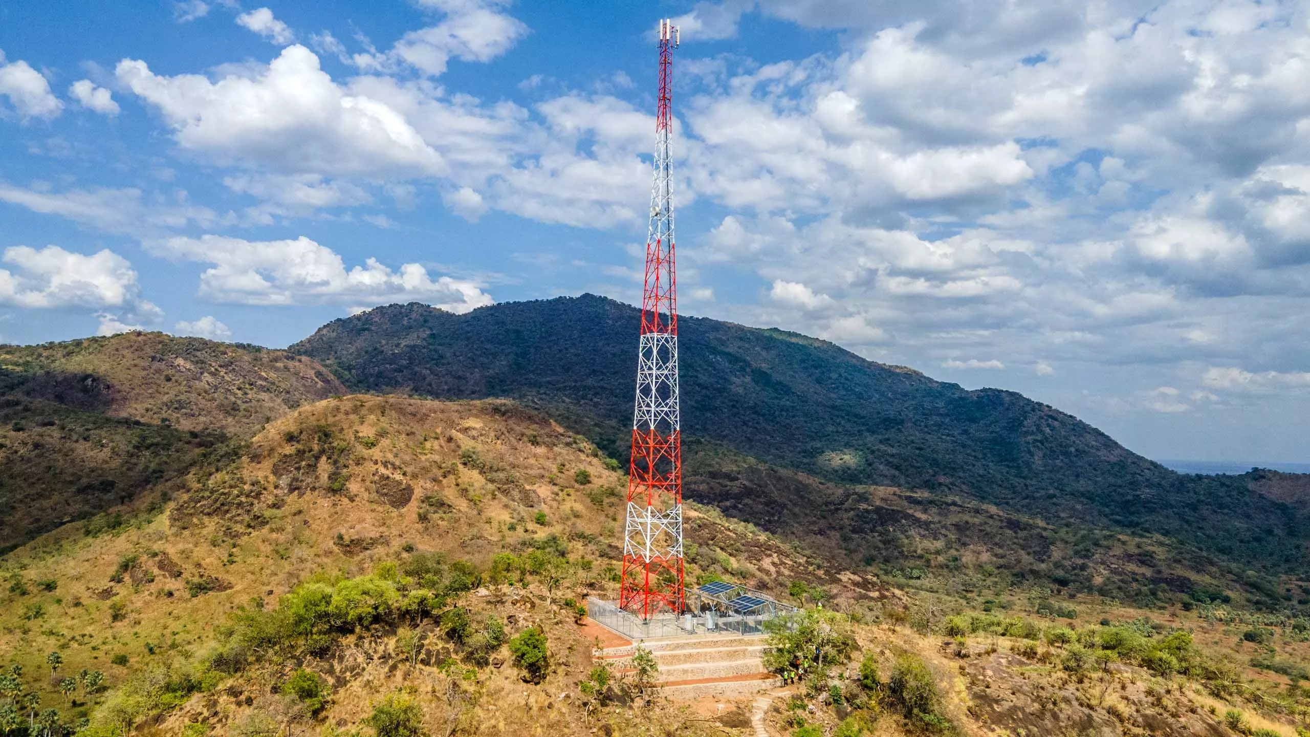 A tall red and white telecommunications tower stands on a hilltop surrounded by mountainous terrain under a partly cloudy sky.