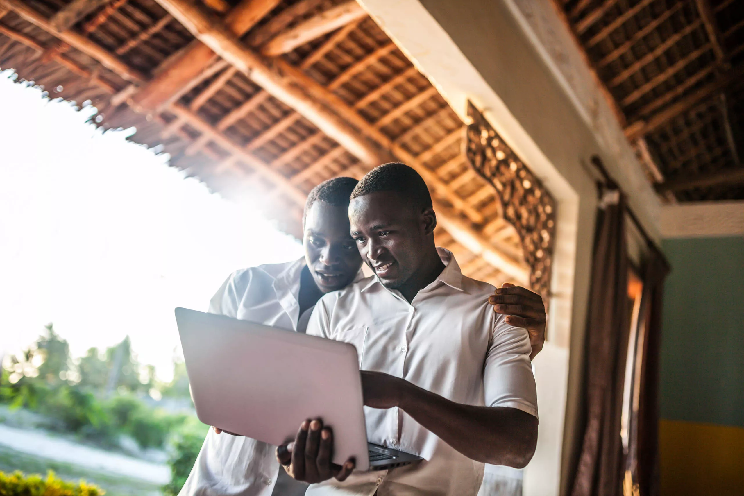 Two people stand close together, smiling and looking at a laptop screen under a wooden roof.