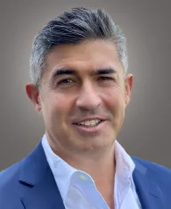 Headshot of a man with a white shirt and suite jacket, on a grey backdrop 